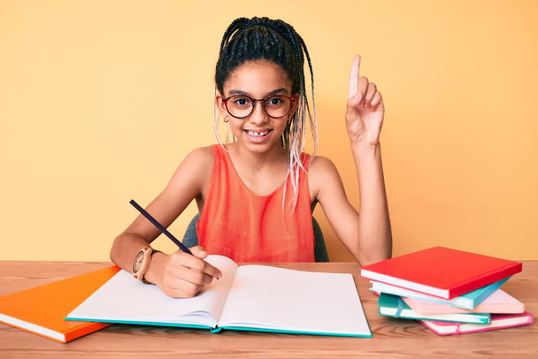 Giovane bambina afroamericana con trecce che studia per l'esame scolastico sorridendo con un'idea o una domanda indicando il dito con la faccia felice, numero uno  - Foto, immagini