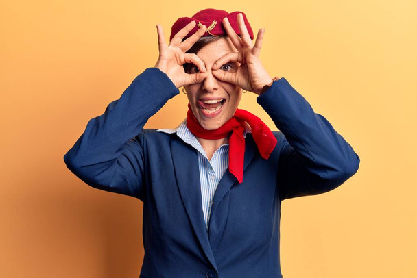 Young beautiful woman wearing stewardess uniform doing ok gesture like binoculars sticking tongue out, eyes looking through fingers. crazy expression.  - Foto, Imagen