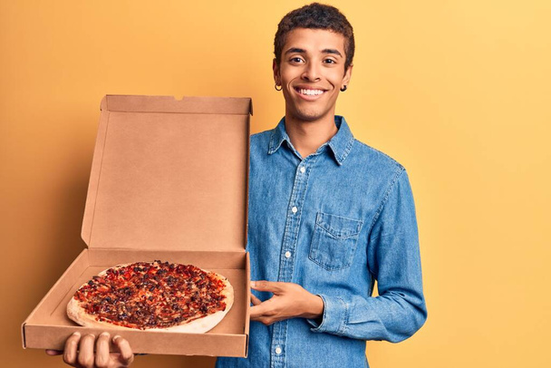 Молодой африканский американец держит доставку пиццы коробка выглядит позитивно и счастливо стоя и улыбаясь с уверенной улыбкой показывая зубы  - Фото, изображение