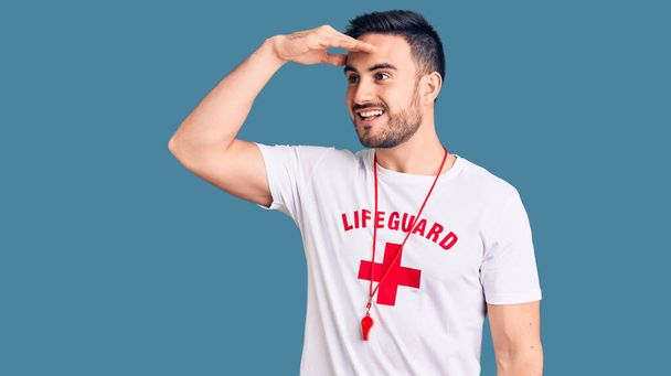 Young handsome man wearing lifeguard uniform very happy and smiling looking far away with hand over head. searching concept.  - Foto, immagini