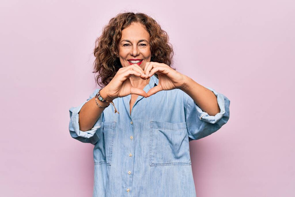 Middelbare leeftijd mooie vrouw dragen casual denim shirt staan over roze achtergrond glimlachend in liefde doen hart symbool vorm met de handen. Romantisch concept. - Foto, afbeelding