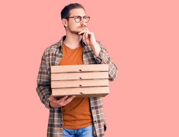 Junge gutaussehende kaukasische Mann mit Lieferung Pizzakiste ernstes Gesicht über Frage mit der Hand auf dem Kinn, nachdenklich über verwirrende Idee  - Foto, Bild