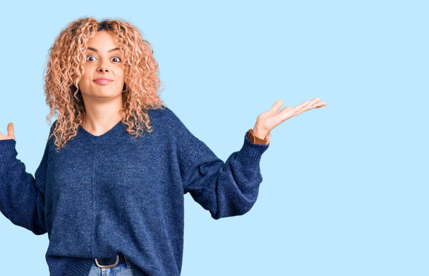Young blonde woman with curly hair wearing casual winter sweater clueless and confused expression with arms and hands raised. doubt concept.  - Photo, Image