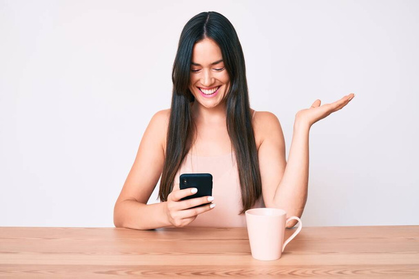 Jeune femme caucasienne assise au bureau en utilisant un smartphone buvant du café célébrant la victoire avec un sourire heureux et l'expression gagnante avec les mains levées  - Photo, image