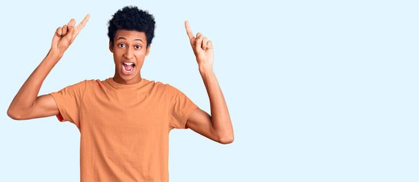 Mladý africký Američan nosí ležérní oblečení, usmívá se ohromeně a překvapeně a ukazuje prstem a zvednutými pažemi.  - Fotografie, Obrázek