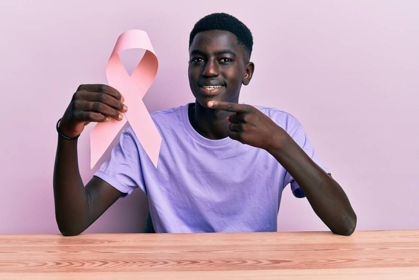 Young african american man holding pink cancer ribbon sitting on the table smiling happy pointing with hand and finger  - Foto, Bild