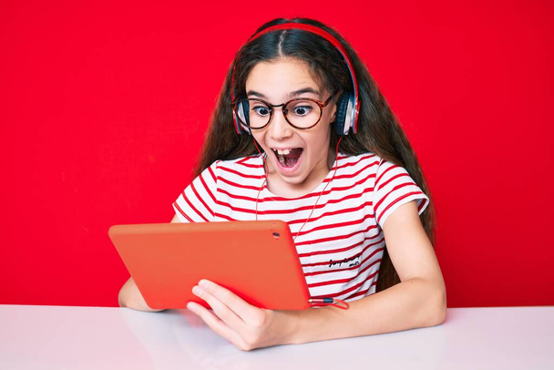 Cute hispanic child girl using touchpad and headphones sitting on the table scared and amazed with open mouth for surprise, disbelief face  - Photo, Image