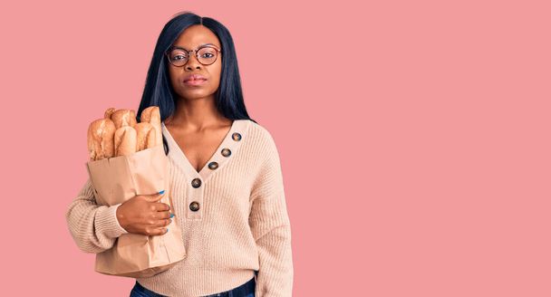 Jonge Afrikaanse Amerikaanse vrouw met papieren zak met brood denken houding en nuchtere uitdrukking op zoek naar zelfvertrouwen  - Foto, afbeelding