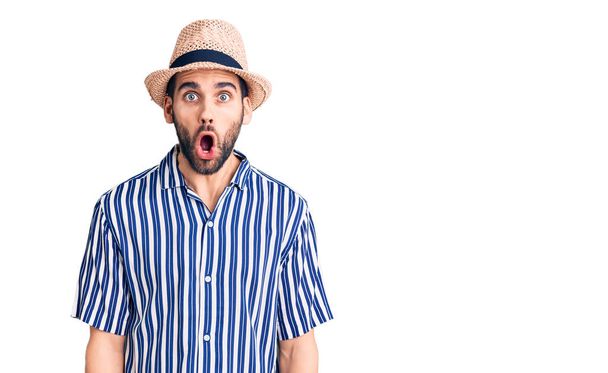 Young handsome man with beard wearing summer hat and striped shirt afraid and shocked with surprise expression, fear and excited face.  - Photo, Image