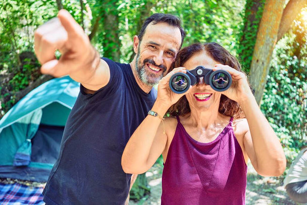 Middle age couple  of hiker camping at the forest. Smiiling happy using binoculars pointing to the side with finger. - Foto, afbeelding