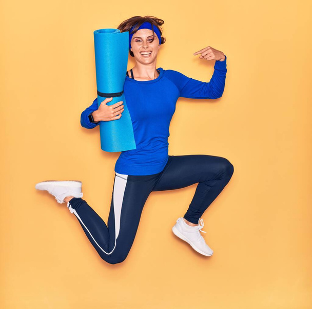 Young beautiful sportswoman smiling happy. Jumping with smile on face holding yoga mat pointing head with finger over isolated yellow background. - Photo, Image