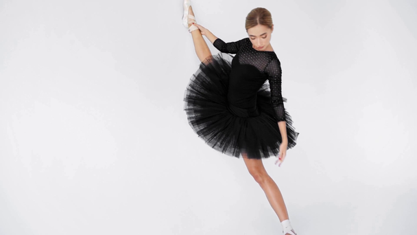 Bailarina joven en tutú negro que estira la pierna cerca de la pared sobre fondo blanco - Imágenes, Vídeo
