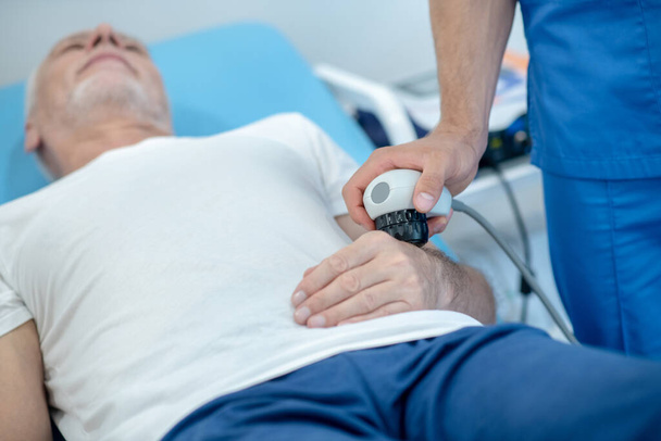 Paziente maschio dai capelli grigi sdraiato sul pullman, sottoposto a trattamento ecografico alle mani - Foto, immagini