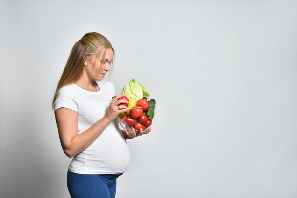 Young pregnant woman holding a bowl of vegetables on a gray background, healthy eating and pregnancy - Foto, afbeelding