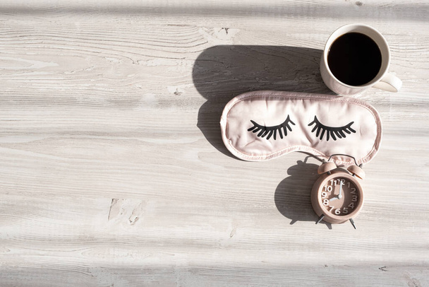 slaapmasker, koffiebeker en wekker op een houten witte tafel in het ochtendzonlicht. Het concept van het starten van een nieuwe dag, goedemorgen of het beëindigen van de dag, laat in de avond. Bedrijvenplanning - Foto, afbeelding