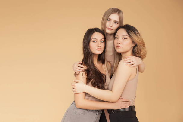 Vrolijke jonge multiculturele Aziatische vrouwen, Kaukasisch knuffelen en lachen in studio op beige achtergrond. Beauty spa behandeling huidverzorging concept - Foto, afbeelding