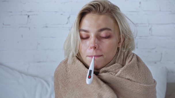 sick woman wrapped in blanket measuring temperature with digital thermometer  - Filmati, video