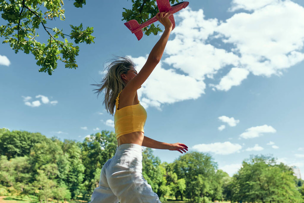 Horizontale Außenaufnahme einer schönen jungen Frau, die im Park läuft, während sie an einem sonnigen Tag mit einem Spielzeugflugzeug spielt. Glückliche Weibchen halten ein Flugzeug bereit, um es zum Fliegen freizugeben. Reisekonzept. - Foto, Bild