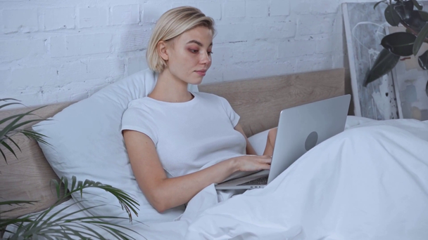молодая блондинка-фрилансер на ноутбуке в спальне  - Кадры, видео