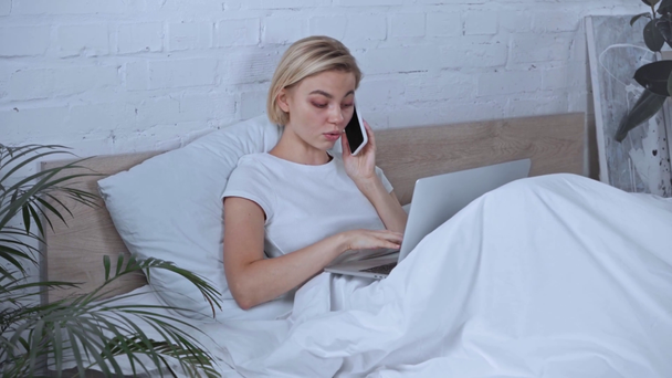 blonde freelancer talking on smartphone near laptop in bedroom - Filmati, video