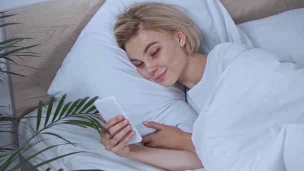 blond vrouw liggend in bed en met behulp van smartphone - Video