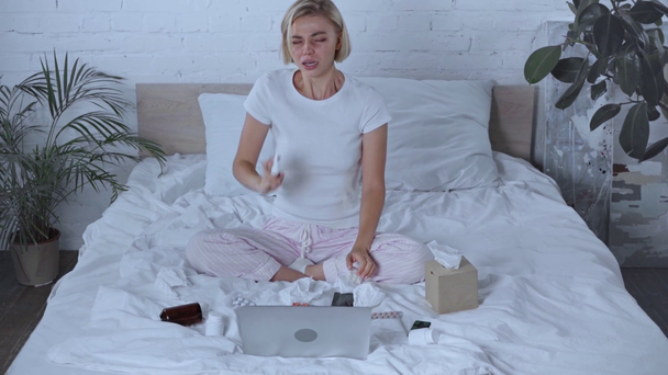 хвора жінка бере серветку і чхає біля ноутбука і смартфона на ліжку
 - Кадри, відео