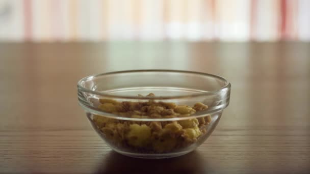 Yoghurt in crunches en cornflakes gieten in een transparante glazen kom om 's ochtends thuis op een houten tafel in het zonlicht te koken. - Video