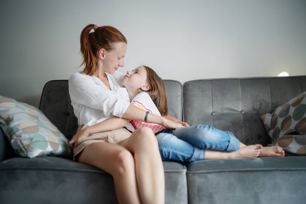 Young beautiful mother and her daughter child 8 years old are sitting on a gray sofa and tenderly hugging, happiness and love in the family. Mother's day concept - Foto, Imagem