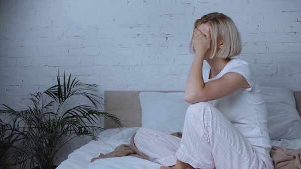 rozrušená blondýna žena sedí na posteli a dotýká tvář  - Záběry, video