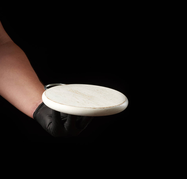 mężczyzna ręka w czarny lateks rękawica posiada okrągły pusty biały drewniany pizza pokładzie, czarne tło - Zdjęcie, obraz