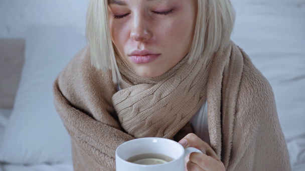 chora młoda kobieta trzyma filiżankę gorącej herbaty i patrzy w kamerę - Materiał filmowy, wideo