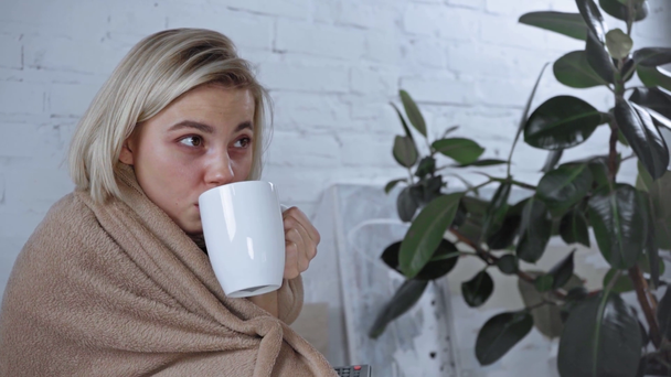 sick young woman holding cup of tea and remote controller while watching film - Footage, Video
