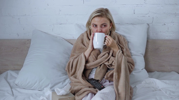 sick woman holding cup of tea, remote controller and watching film in bed - Felvétel, videó