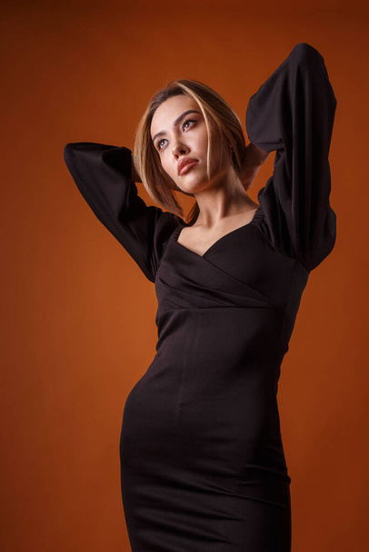elegant model in zwarte jurk met diepe halslijn poserend op oranje achtergrond - Foto, afbeelding