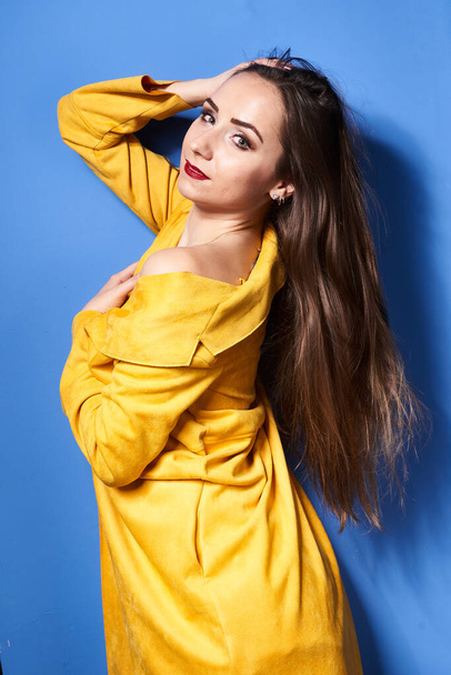  a beautiful girl poses in a yellow coat against a blue wall            - Фото, зображення
