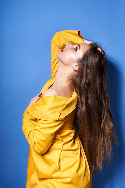  Ένα όμορφο κορίτσι ποζάρει με ένα κίτρινο παλτό στον μπλε τοίχο.            - Φωτογραφία, εικόνα
