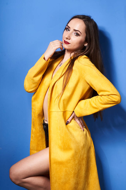  a beautiful girl poses in a yellow coat against a blue wall            - Foto, Imagen