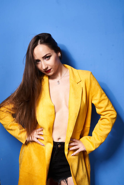  piękna dziewczyna pozuje w żółtym płaszczu na niebieskiej ścianie            - Zdjęcie, obraz