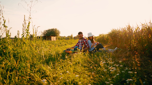casal de agricultores sentados na grama e falando perto da caixa com legumes  - Filmagem, Vídeo