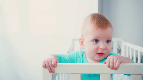 menino de pé no berço do bebê e olhando para a câmera em casa - Filmagem, Vídeo