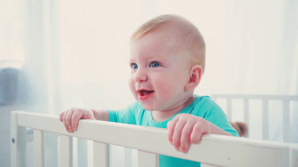 excité bébé garçon debout dans la crèche de bébé et regardant loin - Séquence, vidéo