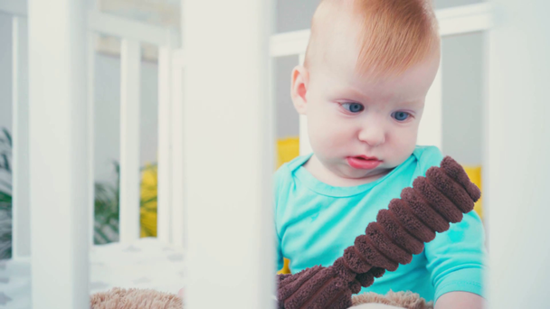 selective focus of infant boy sitting in baby crib with soft toy - Video, Çekim