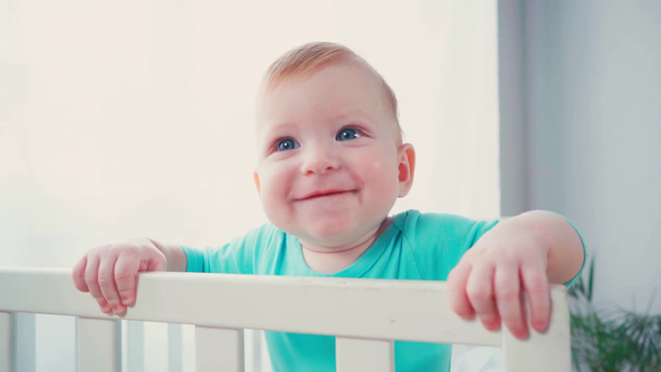 radosny chłopiec stojący w łóżeczku niemowlęcym i patrzący w kamerę - Materiał filmowy, wideo