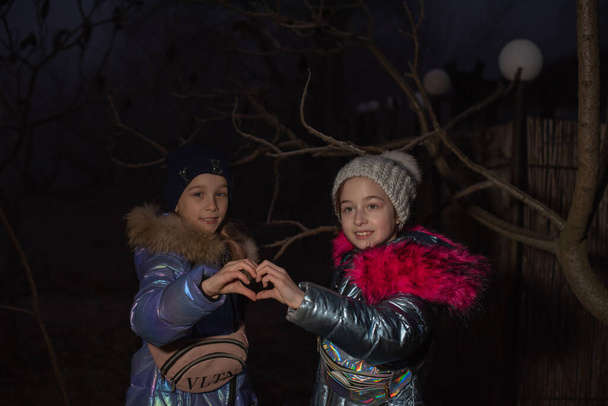 Two little girls are smiling. Happiness. They show the heart with their hands. Two girls in winter jackets show hearts with their hands. Teenagers girls in winter show hearts with their hands. - Photo, Image