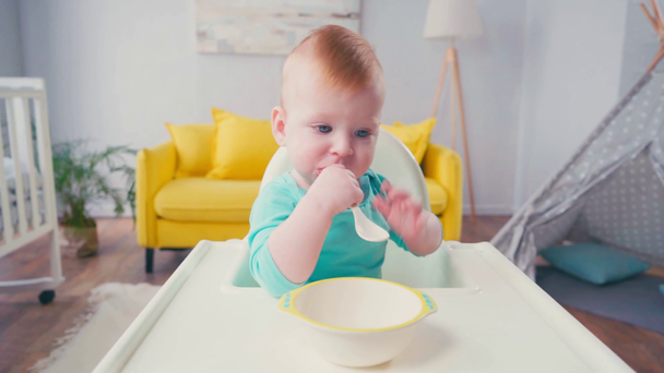bambino seduto sulla sedia del bambino e succhiare cucchiaio vicino ciotola  - Filmati, video