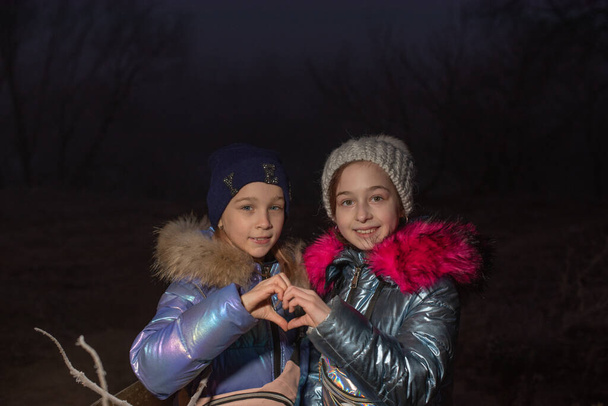 Two little girls are smiling. Happiness. They show the heart with their hands. Two girls in winter jackets show hearts with their hands. Teenagers girls in winter show hearts with their hands. - Photo, Image