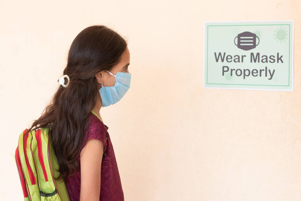 Young girl Student adjusting mask by seeing wear mask properly notice on classroom wall before entering class to protect from coronavirus or covid-19 infection. - Foto, imagen
