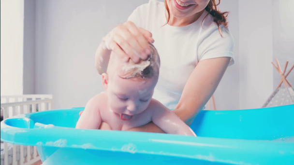 mother washing soapy hair of infant son in baby bathtub  - Footage, Video