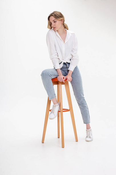 young pensive caucasian woman posing in shirt and blue jeans, sitting on stool - Фото, изображение