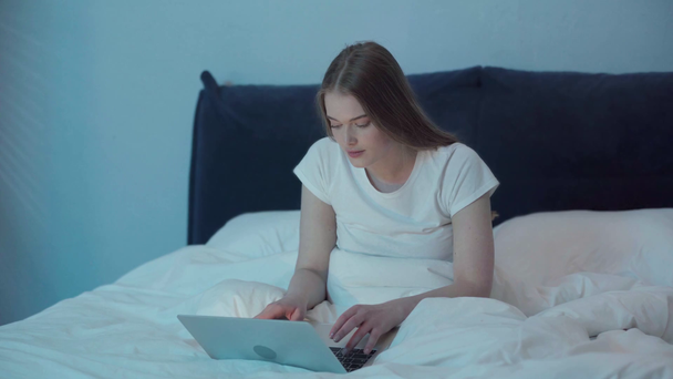 Nuori nainen istuu sängyllä ja kirjoittaa kannettavan yöllä  - Materiaali, video